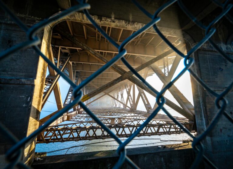 Metal net barrier near modern long suspension stone bridge with metal constructions crossing calm sea on city with bright sunshine